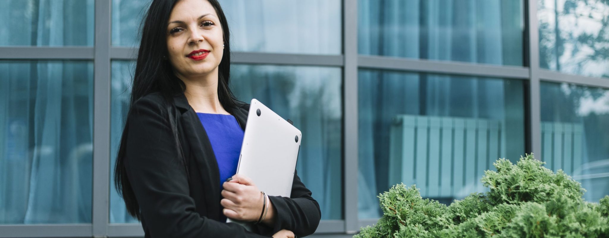 portrait-smiling-businesswoman-holding-laptop-front-glass-building
